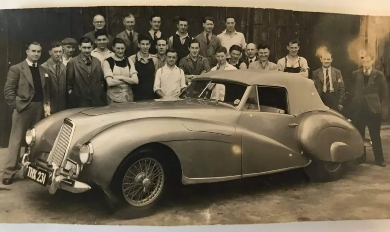 Factory workers standing next to a vintage Aston Martin vehicle