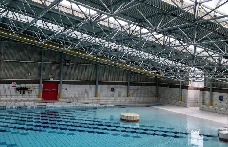 Indoor swimming pool with blue lane markings, tiled walls, and a high metal truss ceiling.