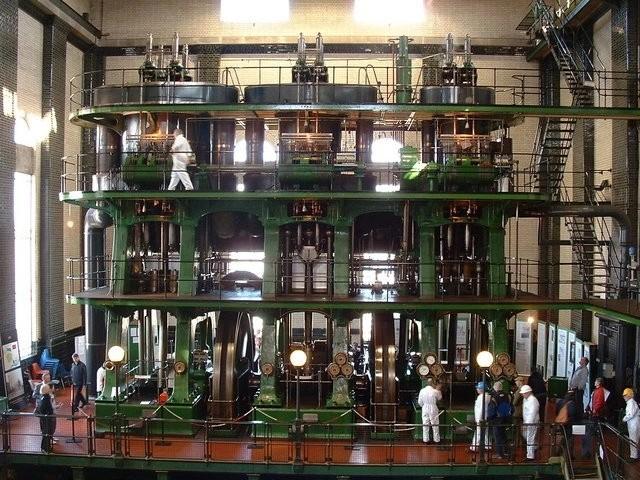 Large green multi-level steam engine with exposed pistons and flywheels inside an industrial hall, with visitors observing.