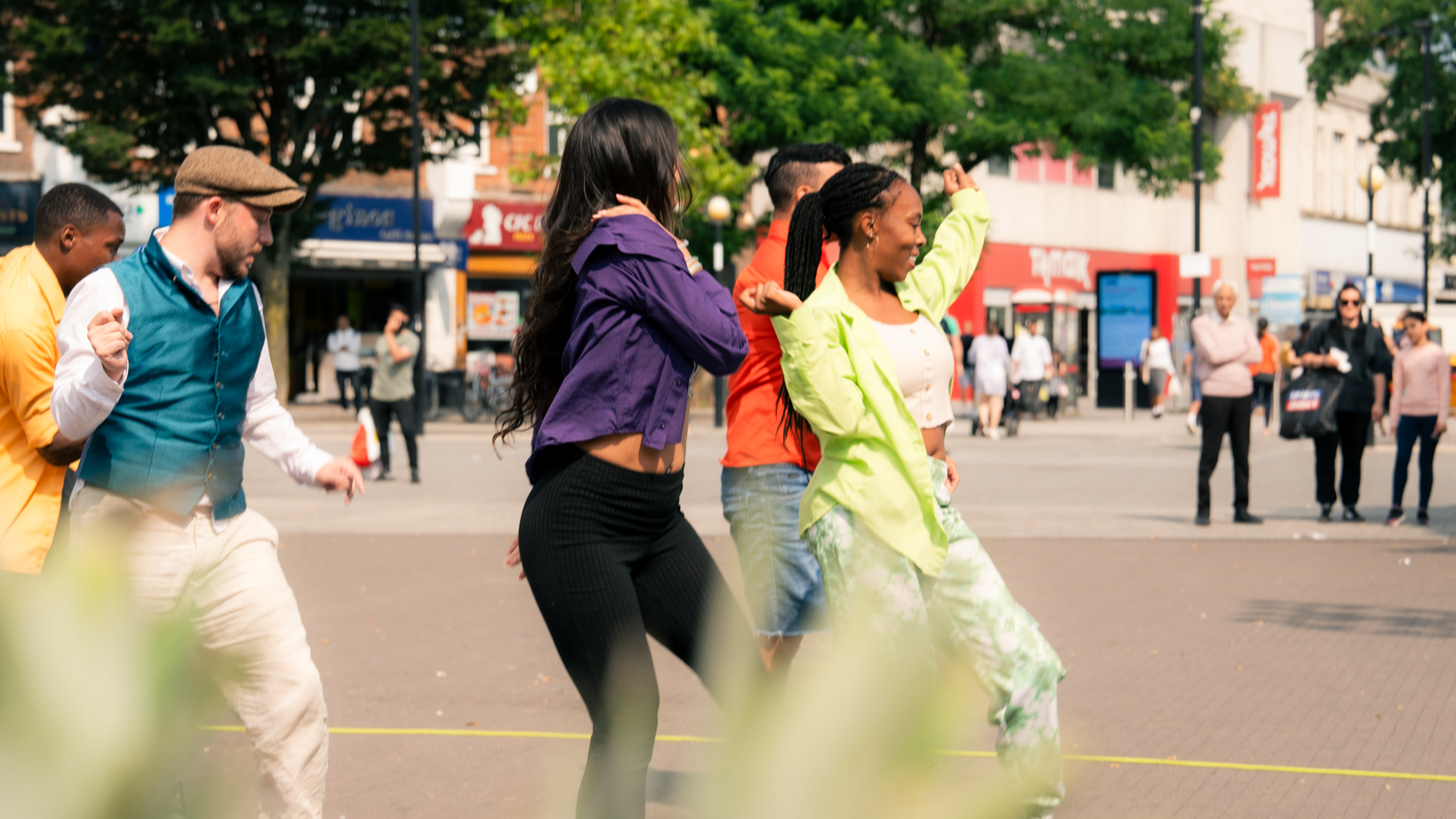 Group of people dancing in an outdoor public square, wearing colourful casual outfits, with shops and trees in the background.