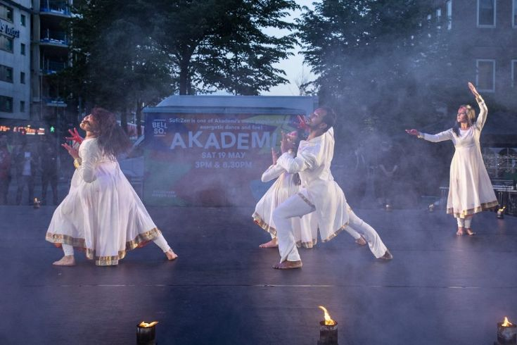 Four dancers in white outfits performing an expressive outdoor routine on a misty stage with lights in Bell Square.